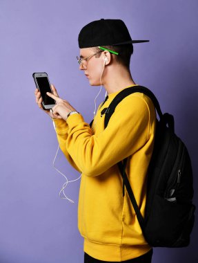 Şapkalı, gözlüklü ve mor arka planda akıllı telefon kullanan neşeli bir öğrencinin portresi.