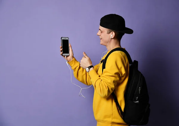 Porträtt av en glad student bär ryggsäck, i mössa och glasögon och använda smartphone över lila bakgrund — Stockfoto