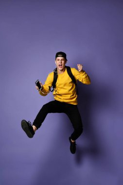 sırt çantası ve kulaklıklar ile gündelik aşınma genç öğrenci adam bir akıllı telefon kullanarak müzik dinliyor ve atlama sırasında gülümseyen