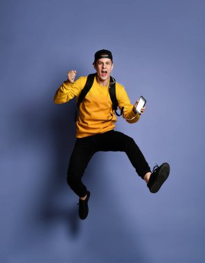 Sırt çantalı ve kulaklıklı yakışıklı genç adam akıllı telefon kullanarak müzik dinliyor ve atlarken gülümsüyor.