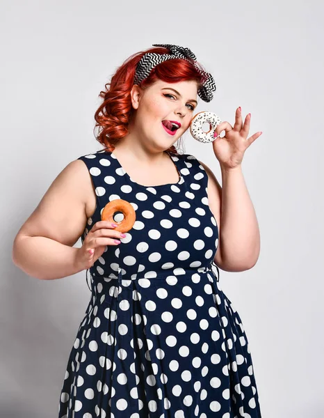 Portret van mooie vrolijke dikke plus size vrouw pin-up dragen van een polka-dot jurk geïsoleerd over lichte achtergrond, het eten van een donut — Stockfoto