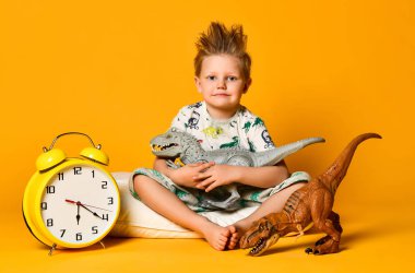 Pijamalı küçük şirin bir çocuk elinde oyuncak bir dinozor tutuyor, elinde çalar saatle yastığın üzerinde oturuyor. Sarı arka planda izole edilmiş..