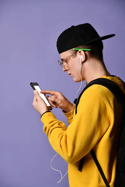 Retrato de um estudante alegre usando mochila, em boné e óculos e usando smartphone sobre fundo roxo — Fotografia de Stock