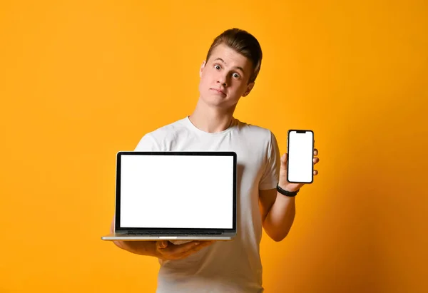 Mladý student muž v bílé košili, drží notebook a mobilní chytrý telefon, stojící ukazuje obrazovku telefonu a počítače v rámečku. — Stock fotografie