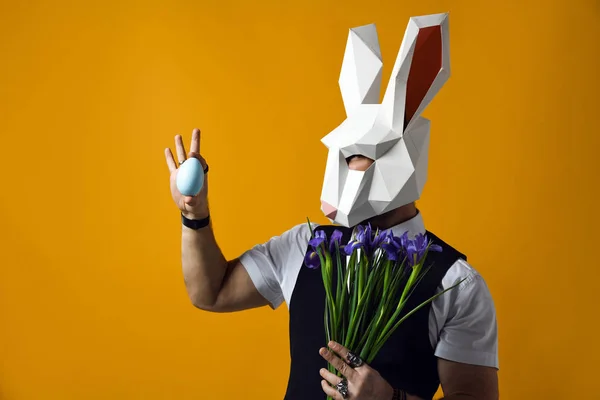 man in paper rabbit mask holds a bouquet of irises flowers on a yellow studio background.