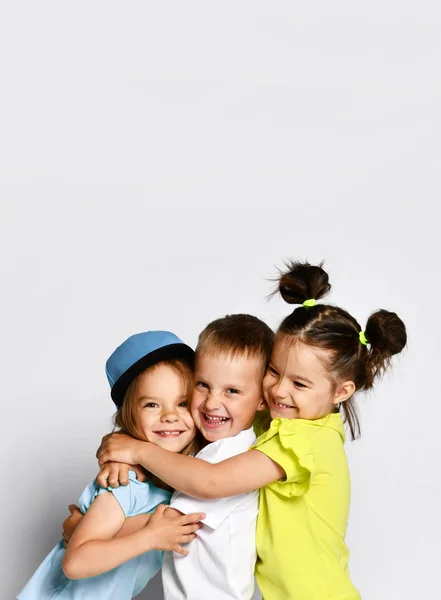 Studio portrait of children on a light background: full body shot of three children in bright clothes, two girls and one boy. Triplets, brother and sisters. hugging on camera. Family ties, friendship — Stock Photo, Image