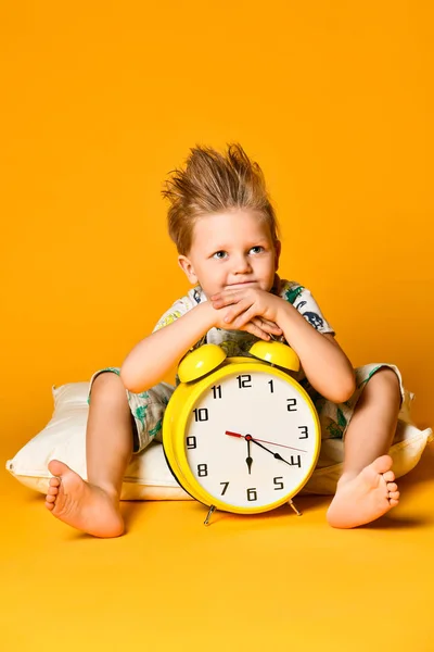 Little cute boy in pajamas holding a toy dinosaur in his hands, sitting on a pillow with an alarm clock. Isolated on a yellow background. Stock Image