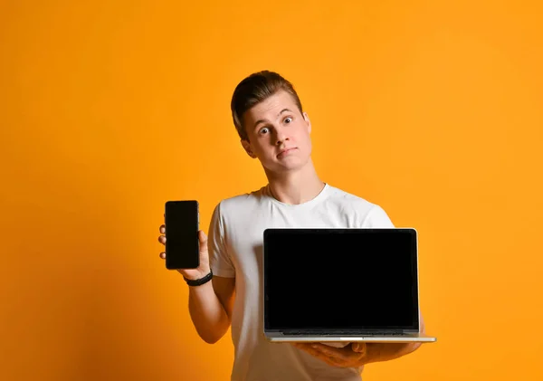 Joven estudiante con una camisa blanca, sosteniendo una computadora portátil y un teléfono inteligente móvil, de pie muestra la pantalla del teléfono y la computadora en el marco . — Foto de Stock