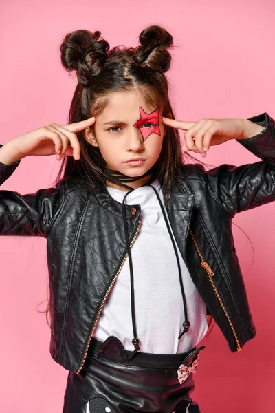 Retrato de una chica punk hipster vestida con una chaqueta y falda de cuero, rodilleras negras y una camiseta blanca, con un peinado divertido y una estrella pintada de maquillaje en la cara, apunta a la cabeza . — Foto de Stock