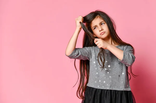 Kleine brunette meisje met lang haar houdt een streng van haar haar en kijkt ernaar. Haarverzorging en kapsel. — Stockfoto
