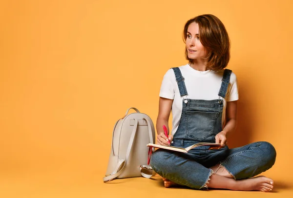 Portrait of a pensive girl in a white T-shirt and denim overalls, sitting on the floor with a red notebook and a pencil on a yellow background — Stock Photo, Image