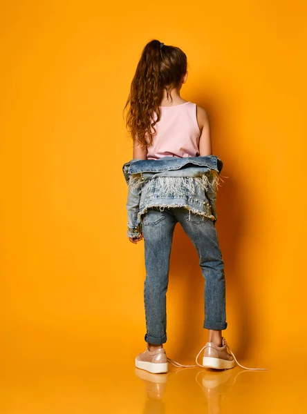 Young teen girl model posing on a yellow background in jeans and a jacket, with long hair gathered in a high tail, stands half a turn to the camera. — Stock Photo, Image