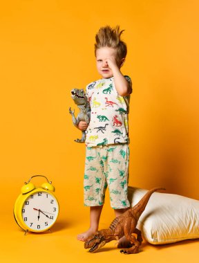 Anaokulu çağında elinde yastıkla ve çalar saatin üstünde uyanan oyuncak bir dinozorla küçük bir çocuk. Stüdyoda sarı arka planda fotoğraf.
