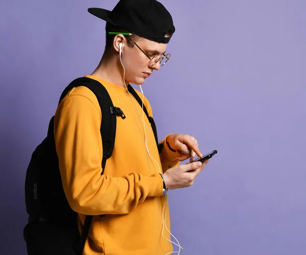 Retrato de um estudante alegre usando mochila, em boné e óculos e usando smartphone sobre fundo roxo — Fotografia de Stock