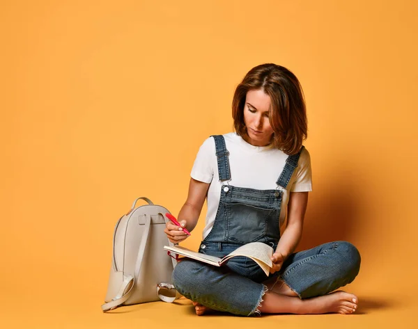 Girl in a white T-shirt and denim overalls, sitting on the floor with a red notebook and a pencil on a yellow background — Stock Photo, Image