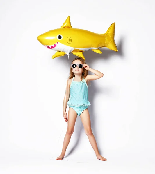 수영복을 입은 긍정적 인 어린 소녀는 노란색 상어 물고기 모양의 풍선을 착용합니다. — 스톡 사진
