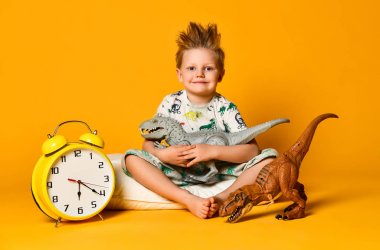 Pijamalı küçük şirin bir çocuk elinde oyuncak bir dinozor tutuyor, elinde çalar saatle yastığın üzerinde oturuyor. Sarı arka planda izole edilmiş..