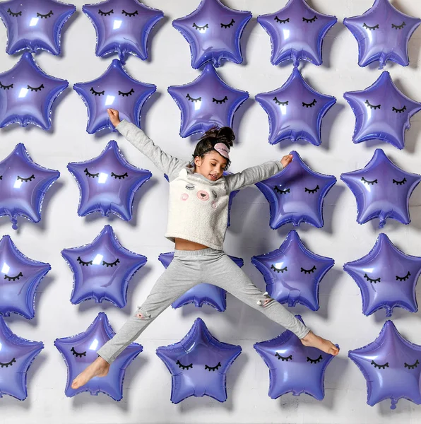 Portrait of a jumping girl in home clothes, happily relaxing together at home and yawning because of insomnia, against the background of balloons in the shape of stars, purple-metallic color — Stock Photo, Image