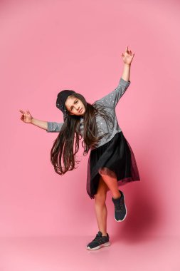 Pembe arka planda, siyah elbiseli, beyzbol şapkalı ve spor ayakkabılı hippi bir kızın portresi.