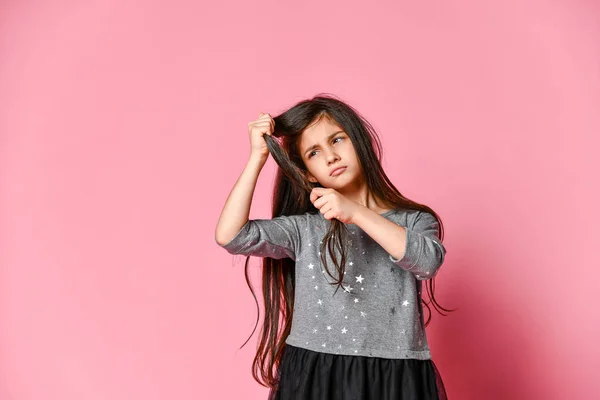 Kleine brunette meisje met lang haar houdt een streng van haar haar en kijkt ernaar. Haarverzorging en kapsel. — Stockfoto