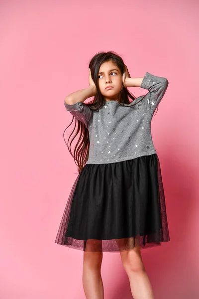 Jong tiener meisje met lang donker haar dragen van een grijze jurk die haar oren met haar handen op een roze achtergrond — Stockfoto