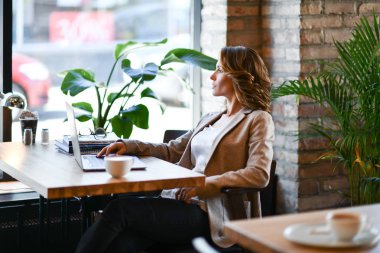 İşine odaklan. Akıllı, rahat giyinen, kendine güveni tam genç bir kadın kreatif ofis veya kafenin penceresinin yanında otururken dizüstü bilgisayarla çalışıyor.