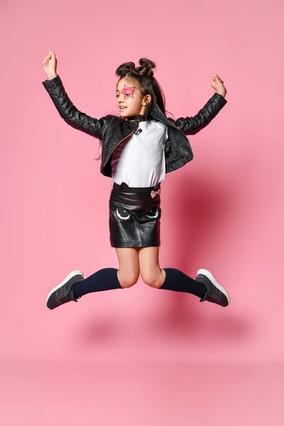 Modieuze hipster punk Girl-gekleed in een lederen jas en rok, zwarte knie-highs en een wit t-shirt, sprongen hoog op een roze — Stockfoto