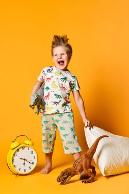 Anaokulu çağında elinde yastıkla ve çalar saatin üstünde uyanan oyuncak bir dinozorla küçük bir çocuk. Stüdyoda sarı arka planda fotoğraf.