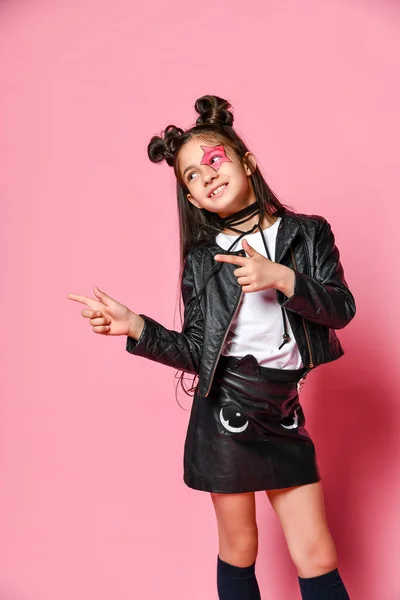 Little - modieuze hipster punk meisje - gekleed in een leren jas en rok, zwarte knie-highs en een wit T-shirt, met een grappig kapsel en een make-up ster op het gezicht, wijst naar de zijkant — Stockfoto