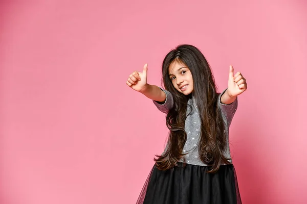 Retrato de una niña muy atractiva con un vestido oscuro, mostrando los pulgares hacia arriba con dos manos sobre un fondo rosa — Foto de Stock