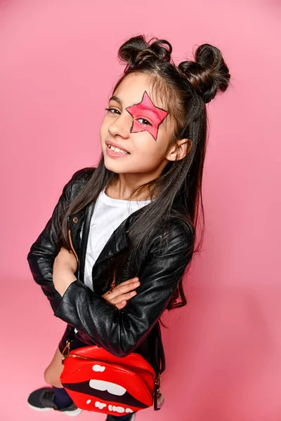 Pequeña chica hipster punk de moda vestida con una chaqueta y falda de cuero, loaches negros y una camiseta blanca, con un peinado divertido y una estrella de maquillaje pintada en su cara. — Foto de Stock