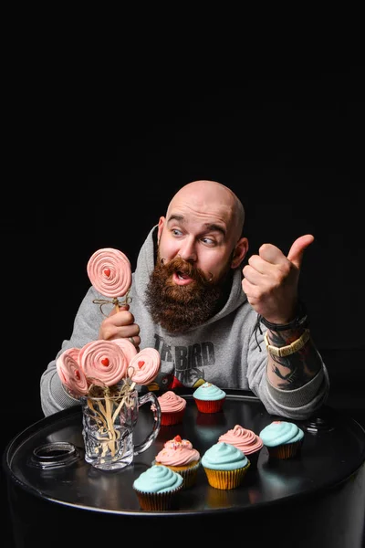 Happy bearded bald man holding two cream cakes on black background.