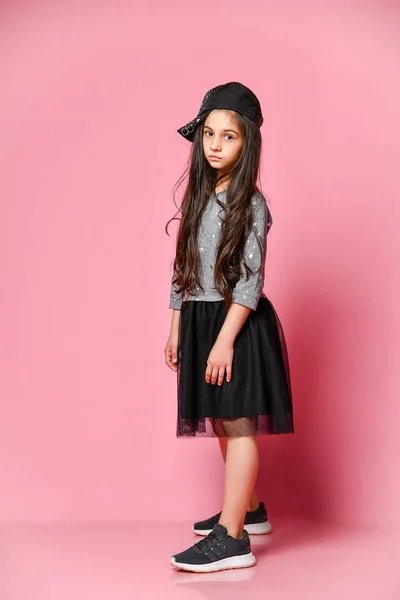 Portrait of a little hipster girl in a dark dress with a baseball cap and sneakers, on a pink background