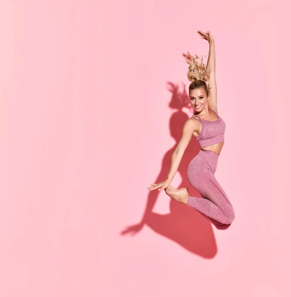 Happy Athletic kobieta skoki w sylwetki. Zdjęcie sportowej kobiety w modnej różowej odzieży sportowej na różowym — Zdjęcie stockowe