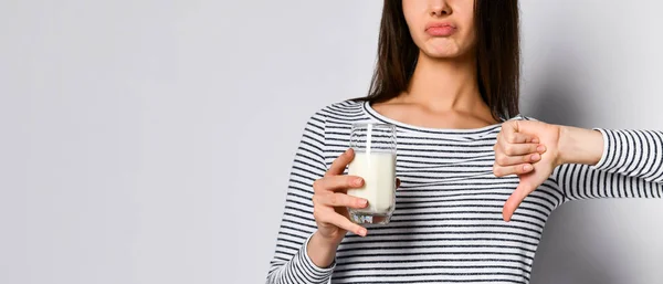 Mujer joven disgustada sosteniendo un vaso con leche de vaca y mostrando un mal pulgar abajo — Foto de Stock