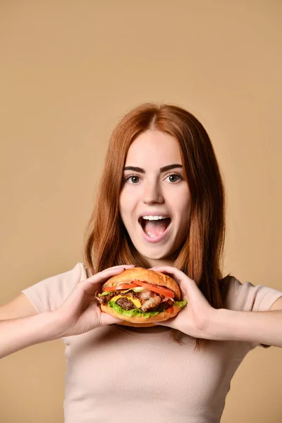 Young red-haired girl holds a burger sandwich, is surprised, on a beige background — Stock Photo, Image