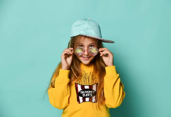 Pretty fashionable young teenager with a lovely smile posing in a baseball cap and yellow hoodie on a blue background — Stock Photo, Image