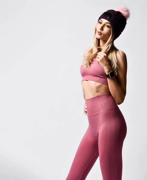 Fit mujer deportiva se ve en ropa deportiva rosa y un sombrero de punto con un pompón mira su reloj inteligente —  Fotos de Stock