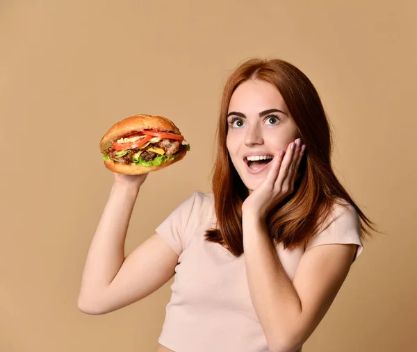 Jeune fille rousse tient un sandwich burger, est surprise, sur un fond beige — Photo