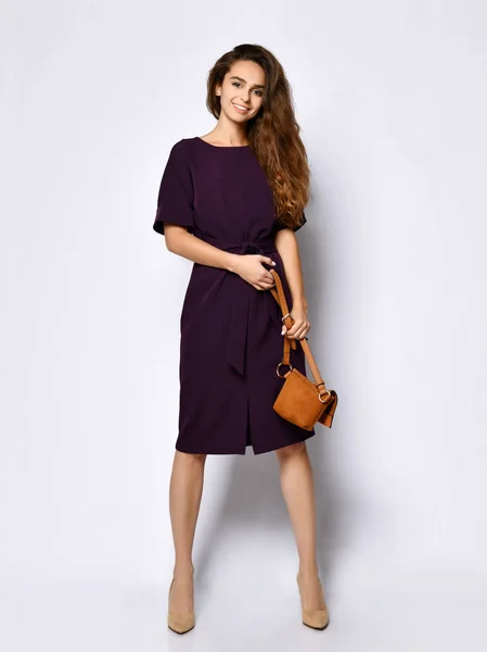 Modelo feminino encaracolado esbelto em um vestido longo escuro roxo e uma pequena bolsa marrom, em pleno crescimento . — Fotografia de Stock
