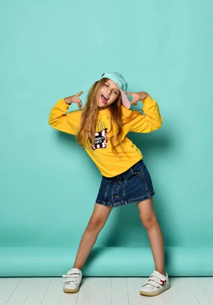 Beyzbol şapkalı ve sarı kapüşonlu komik genç kız mavi arka planda oynaşıyor.. 