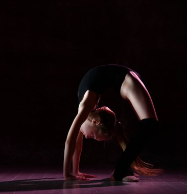 Siyah spor vücutlu ve özel ayakkabılı genç kız jimnastikçi briç pozisyonunda duruyor ve koyu arkaplanda dengesini koruyor.