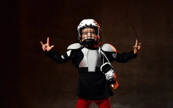 Pozytywny chłopiec w mundurze hokejowym i kasku ochronnym z łyżwami hokejowymi stojącymi i pokazującymi dobry znak kciuka — Zdjęcie stockowe