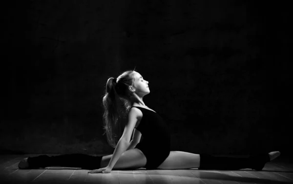 Молодая улыбающаяся девушка гимнастка с длинными волосами в черном спортивном теле и верхние сидят в бечевке боком на темном фоне — стоковое фото