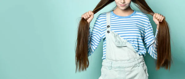 Teen lady in jeans overall and striped sweatshirt. She is holding her long hair, posing against blue studio background. Close up — Stock Photo, Image