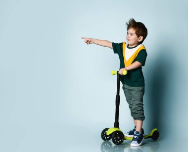 Renkli tişörtlü çocuk, gri kot pantolon, spor ayakkabı. Bir şeyi işaret ediyor, mavi arka planda scooter 'ın üzerinde duruyor.