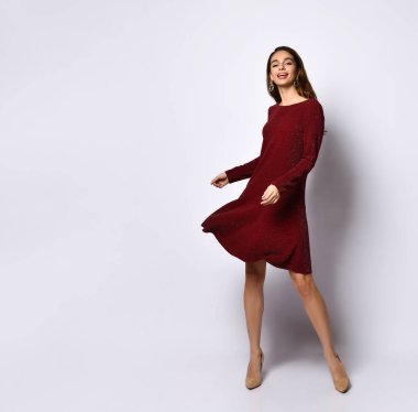 Şirin, zayıf, genç, gülümseyen bir model kısa koyu kırmızı pul elbise ve pastel topuklu ayakkabılar gösteriyor. Beyaz üzerine izole edilmiş tam boy portre