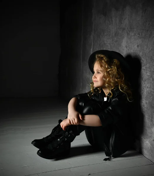 Retrato de menina em terno de couro preto e chapéu no cabelo dourado sentado em holofotes perto da parede escura abraçando seus joelhos — Fotografia de Stock