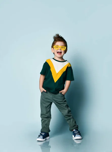 Niño en camiseta colorida, gafas de sol amarillas, jeans grises, zapatillas de deporte. Manos en los bolsillos, boca abierta, posando sobre fondo azul — Foto de Stock