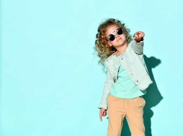 Cute little curly girl with black non-transparent glasses on face stands raising one hand pointing two fingers up. Studio shot isolated on turquoise